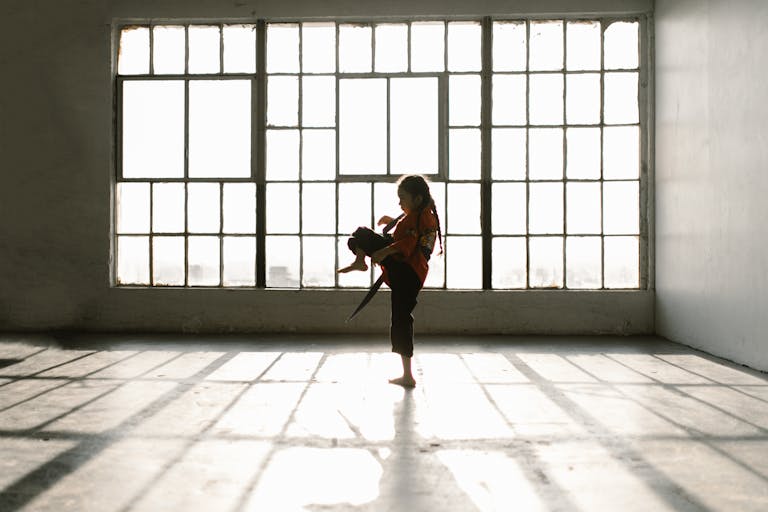 A young girl performs a karate kick in a sunlit room, casting dramatic shadows.