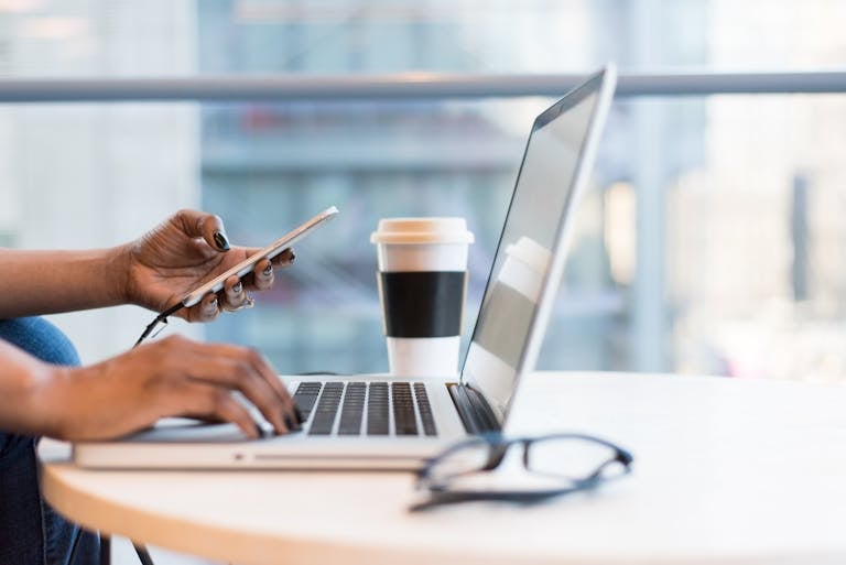 Close-up of hands using a laptop and phone with coffee on a modern office desk.