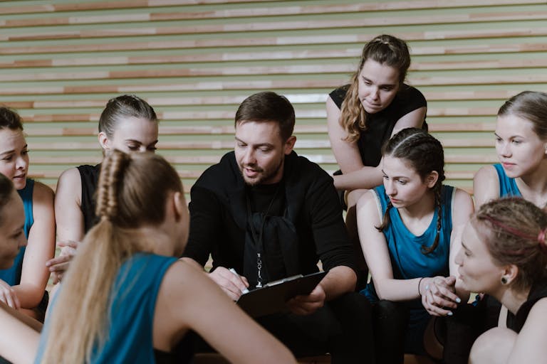 A coach discusses strategy with a women's team seated in a gym setting.