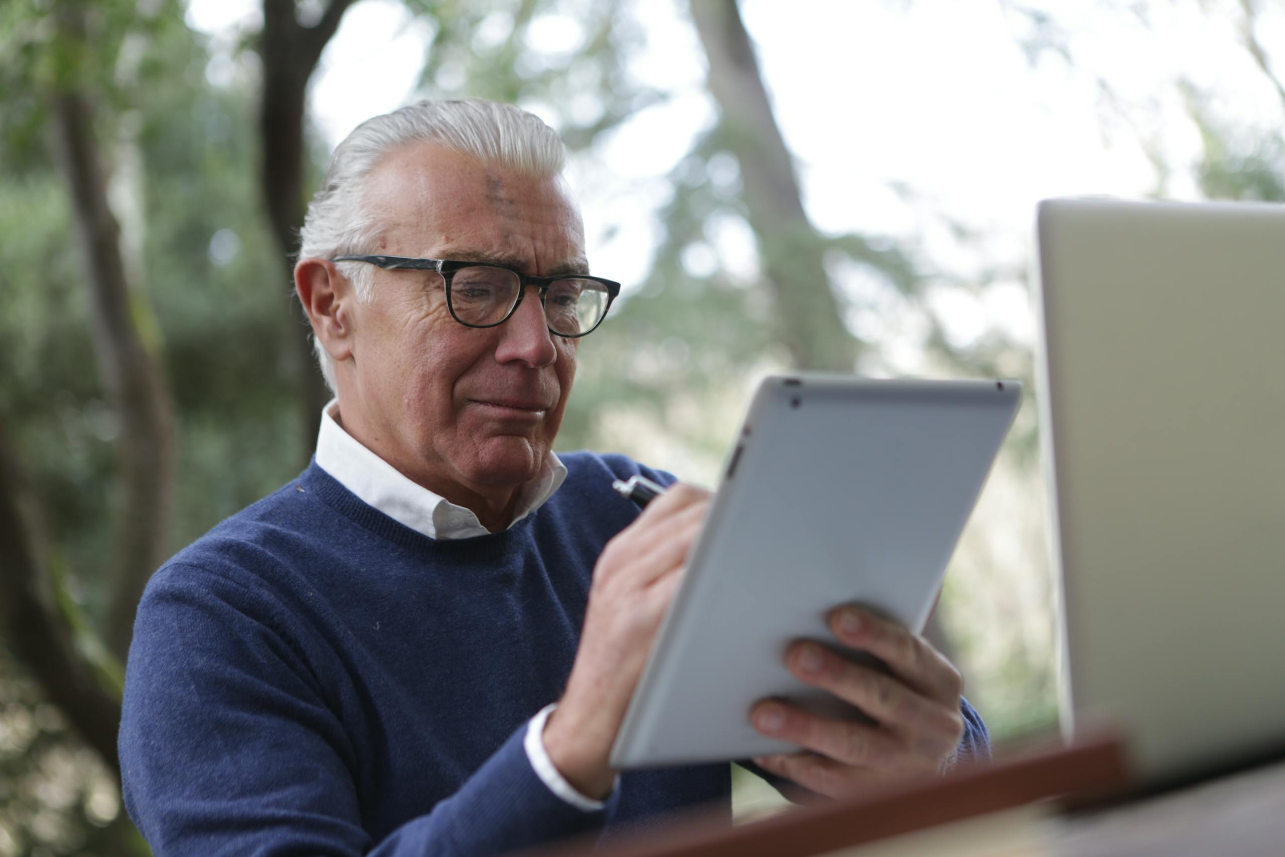 Elderly man using a tablet and laptop in an outdoor setting, indicating modern senior lifestyle.