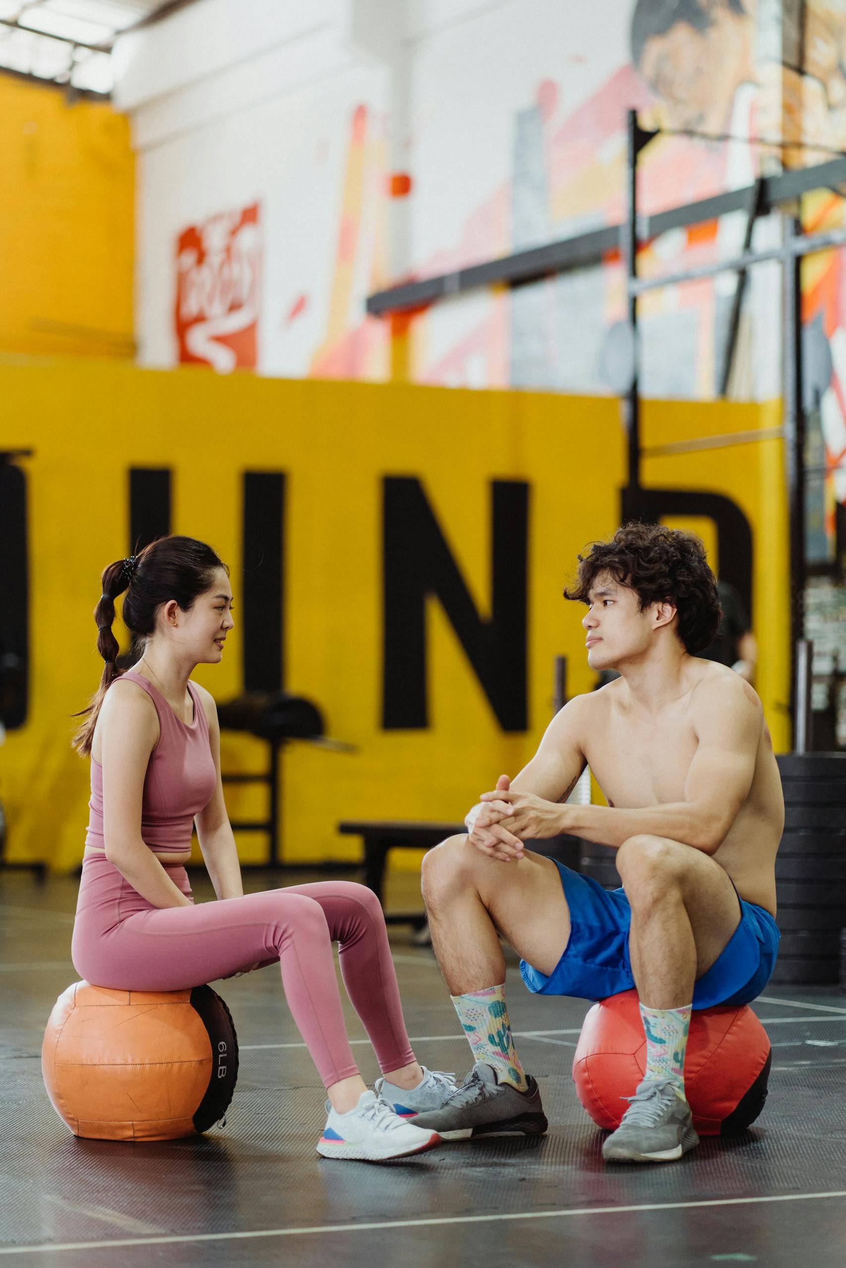 A young man and woman resting and chatting while sitting on medicine balls in a vibrant gym setting.